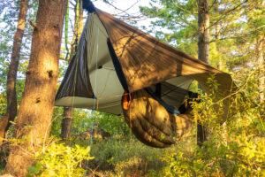 A suspended tree tent setup in a forest, anchored between sturdy trees with wide straps. The tent includes an insulated sleeping bag and rainfly.