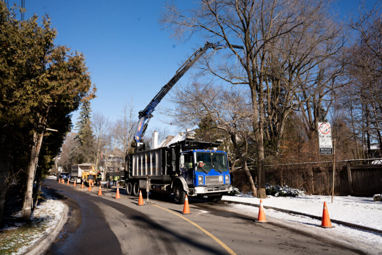 Arborists working with a crane and trucks to clear tree debris along a snowy road.