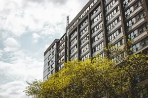 A brown apartment building with a green tree in the foreground and blue sky with white clouds in the background.
